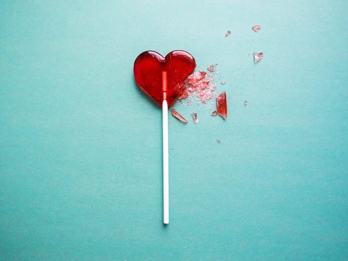 A red, heart-shaped lollipop with part of it broken and shattered pieces scattered on a teal background.
