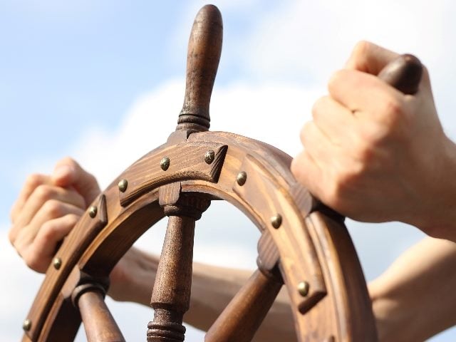 Hands guide a ship's wheel against a blue sky with white clouds in piece about data strategists. 