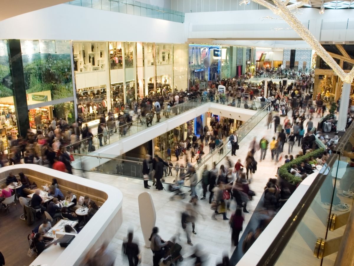 A busy shopping mall with multiple levels crowded with shoppers walking, browsing stores, and dining in a food court.