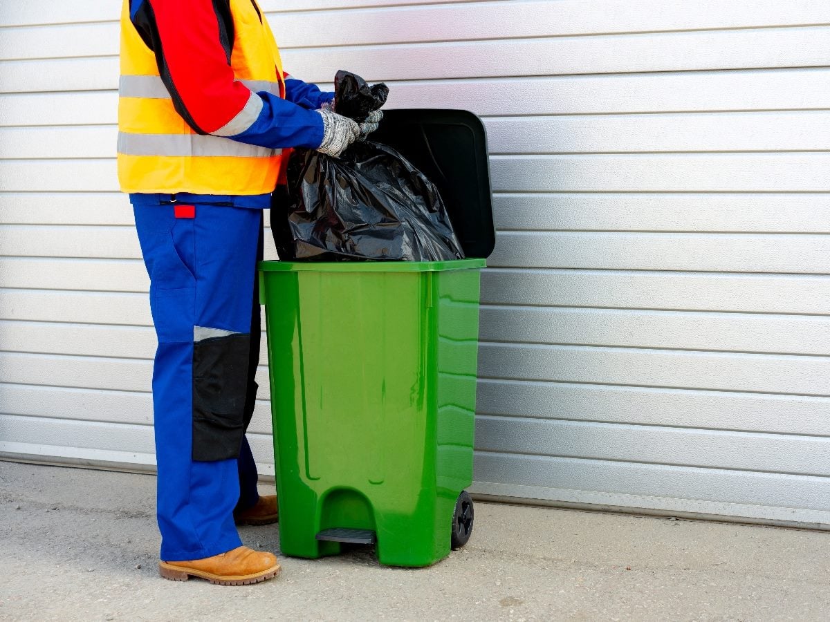 Worker placing a black trash bag into a green outdoor garbage bin.