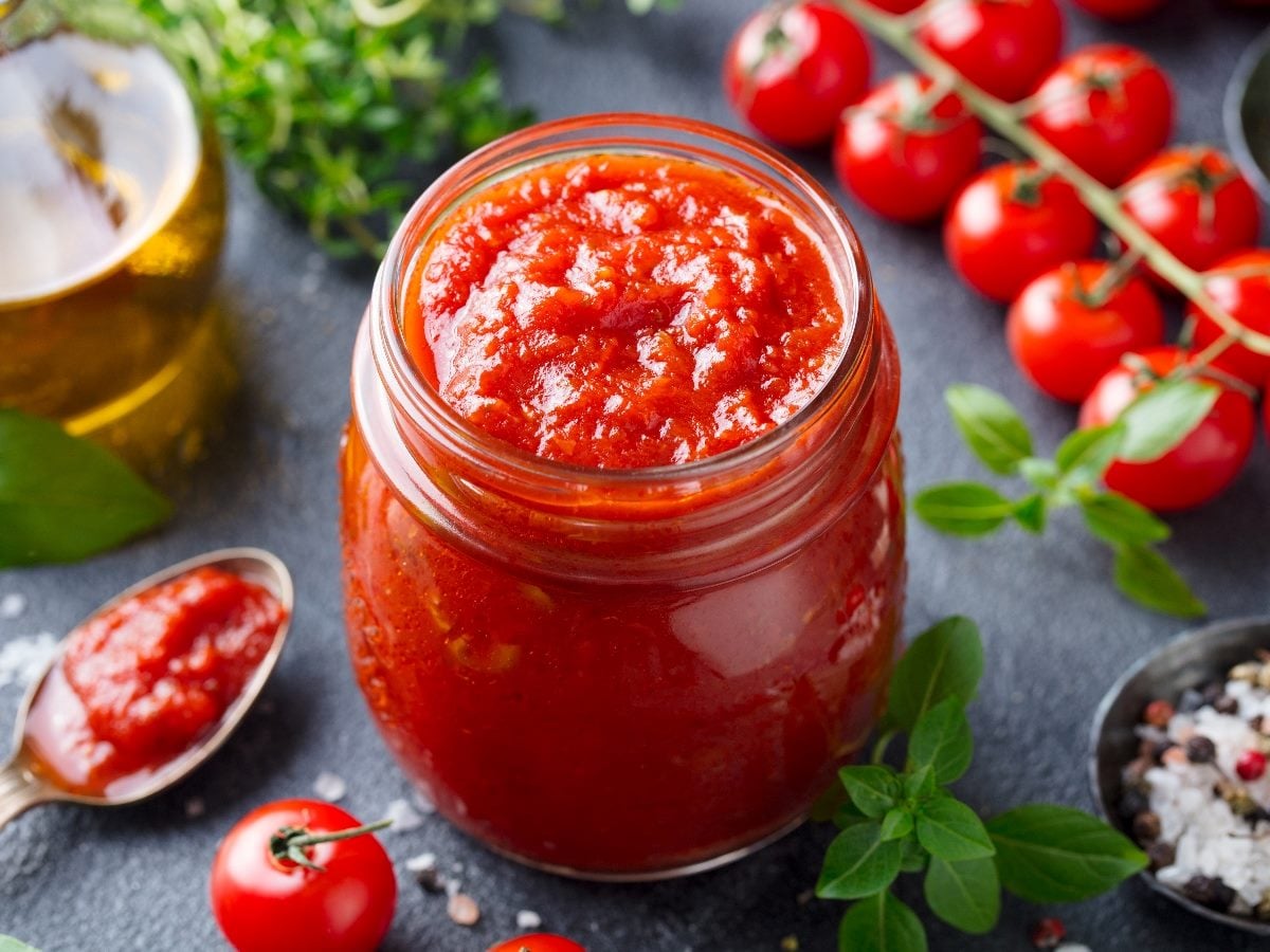 Jar of homemade tomato pasta sauce surrounded by fresh cherry tomatoes, basil leaves, olive oil and seasonings on a dark countertop, highlighting fresh ingredients commonly used in Italian cooking.