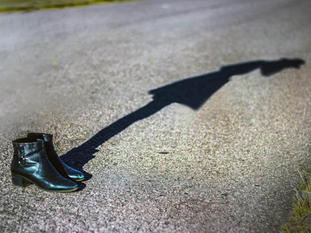 A pair of black boots on a paved road, casting a long shadow stretching forward.