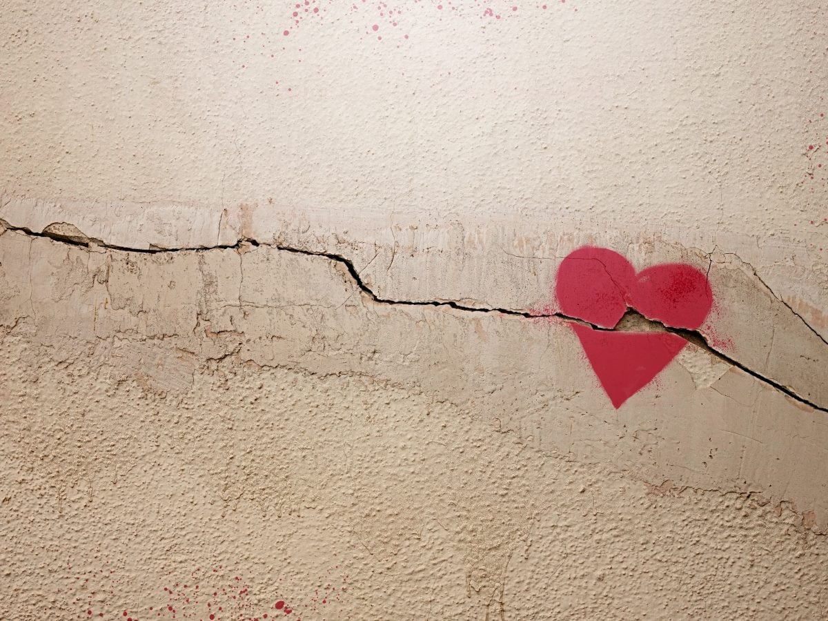 A red heart stenciled in spray paint sits directly over a long horizontal crack running across a textured beige plaster wall, with the crack visibly splitting the heart in two, and faint pink paint spatter scattered across the upper and lower portions of the wall's rough surface.