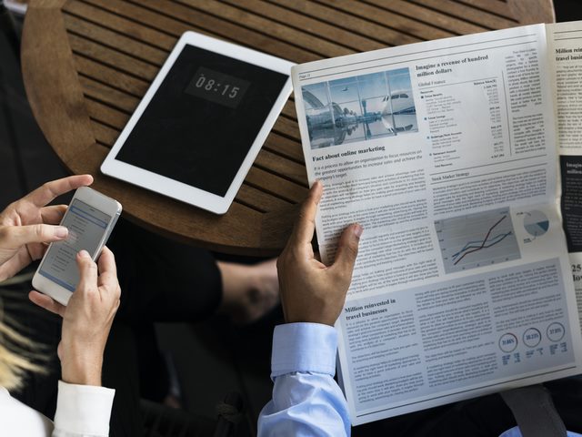 A woman's hands holding a smartphone, and a man's hands holding a newspaper.