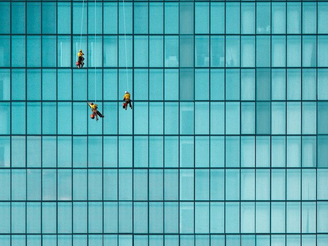 three window washers against a glass covered building,  suspended  high above the street