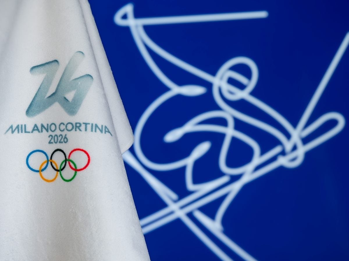 White fabric featuring the Milano Cortina 2026 logo and Olympic rings in the foreground, set against a blue background with a stylized line illustration of a skier in motion.