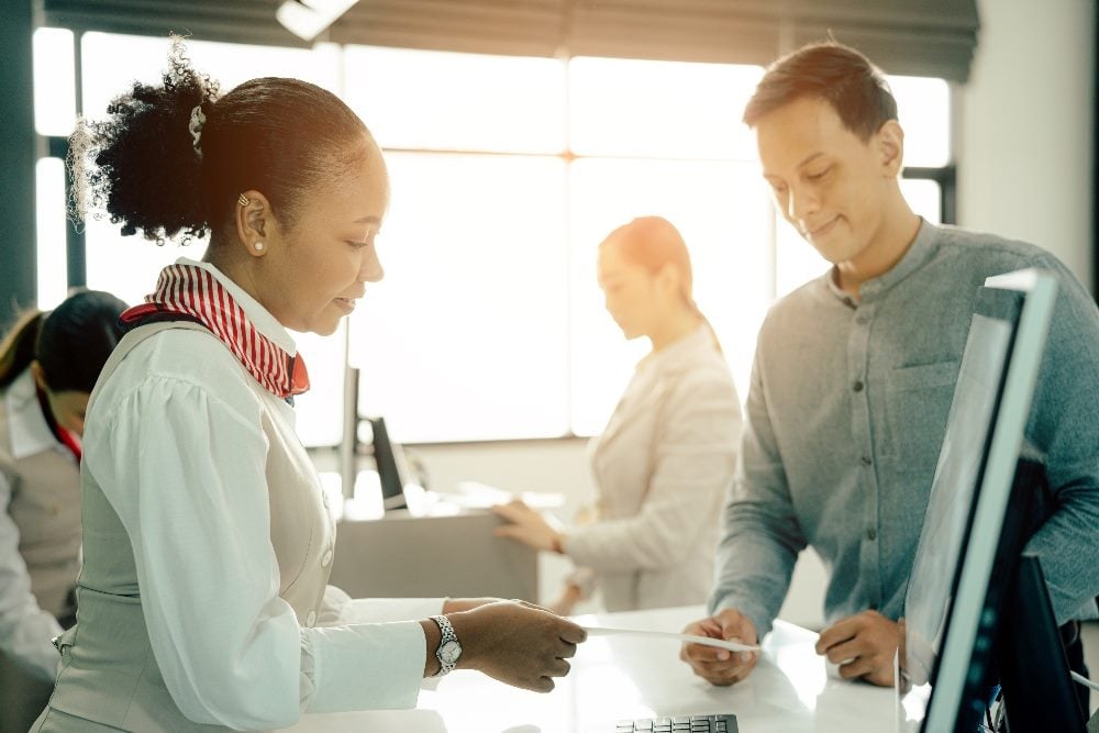 An airline employee at the airport checks the boarding pass for a customer with another customer and employee and large window visible in the blurred background in a piece about America Airlines customers service and automated customer service.