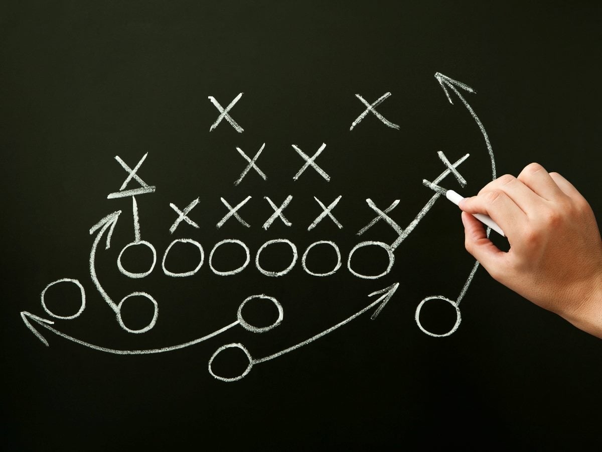 A hand draws a football play on a chalkboard, showing Xs, Os and directional arrows mapping multiple routes toward a successful outcome. The image symbolizes strategic planning, coordinated execution and following a structured playbook to achieve a goal.