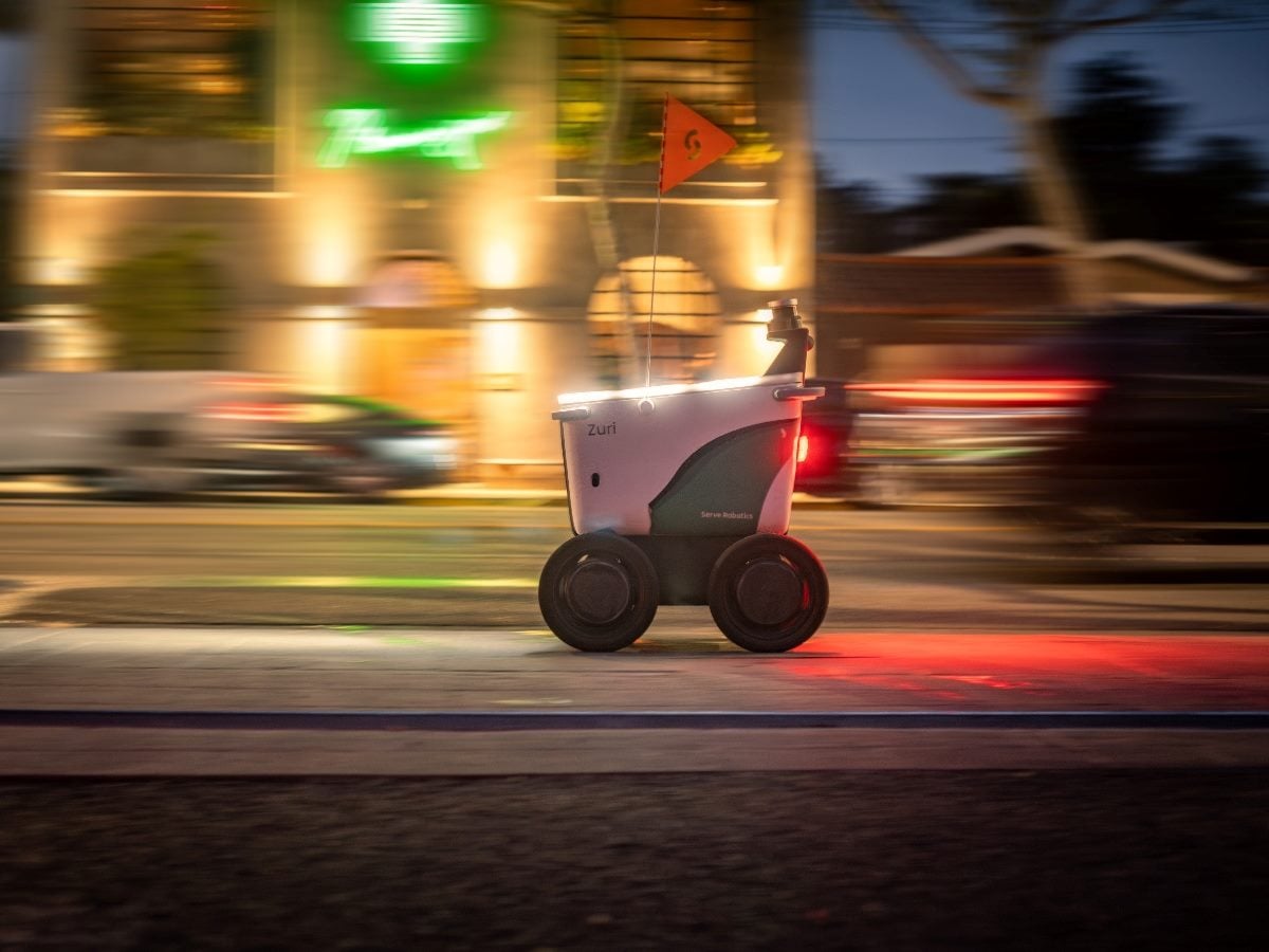 A small autonomous delivery robot travels along a city street at dusk, illuminated by surrounding lights and vehicle headlights. Motion blur in the background emphasizes movement as the four-wheeled robot, carrying a small orange safety flag, navigates an urban environment independently.