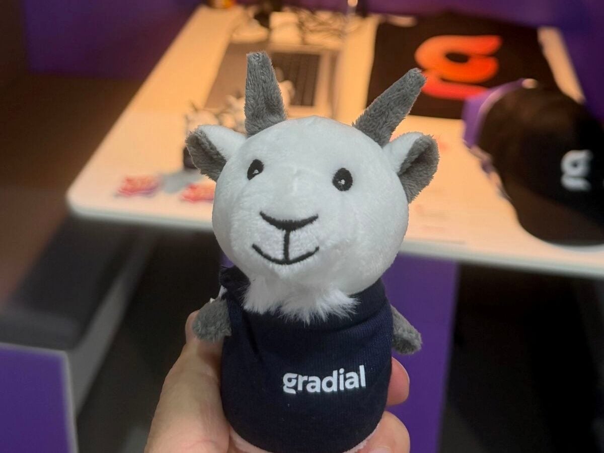 A hand holds a small plush goat mascot wearing a navy-blue hoodie with the Gradial logo. The stuffed animal is centered in the foreground, with a blurred trade show booth in the background featuring purple lighting, a white table and branded promotional items.
