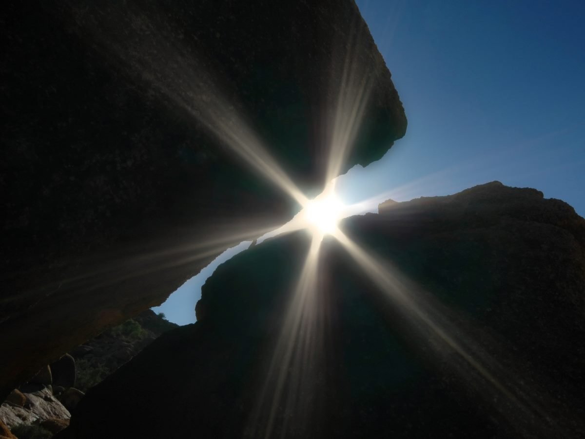 Sunlight bursting through a narrow gap between two large rock formations, creating bright starburst rays against a clear blue sky.