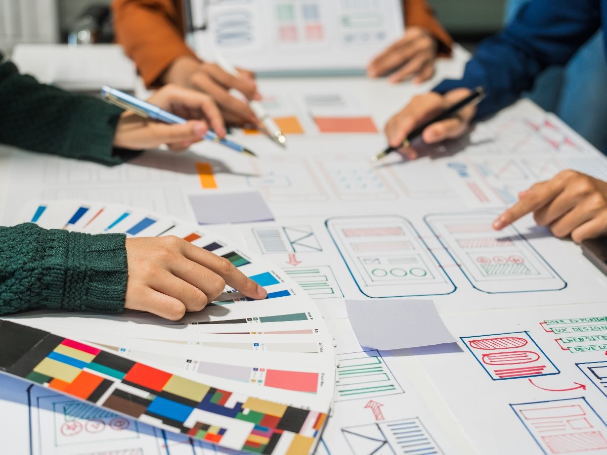 A group of people, showing just their hands, collaborate around a desk, discussing and reviewing user interface (UI) and user experience (UX) elements.