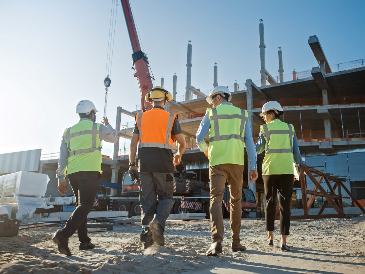 Four construction professionals wearing hard hats and high-visibility vests walk toward a building under construction, with a crane and structural framework in the background.