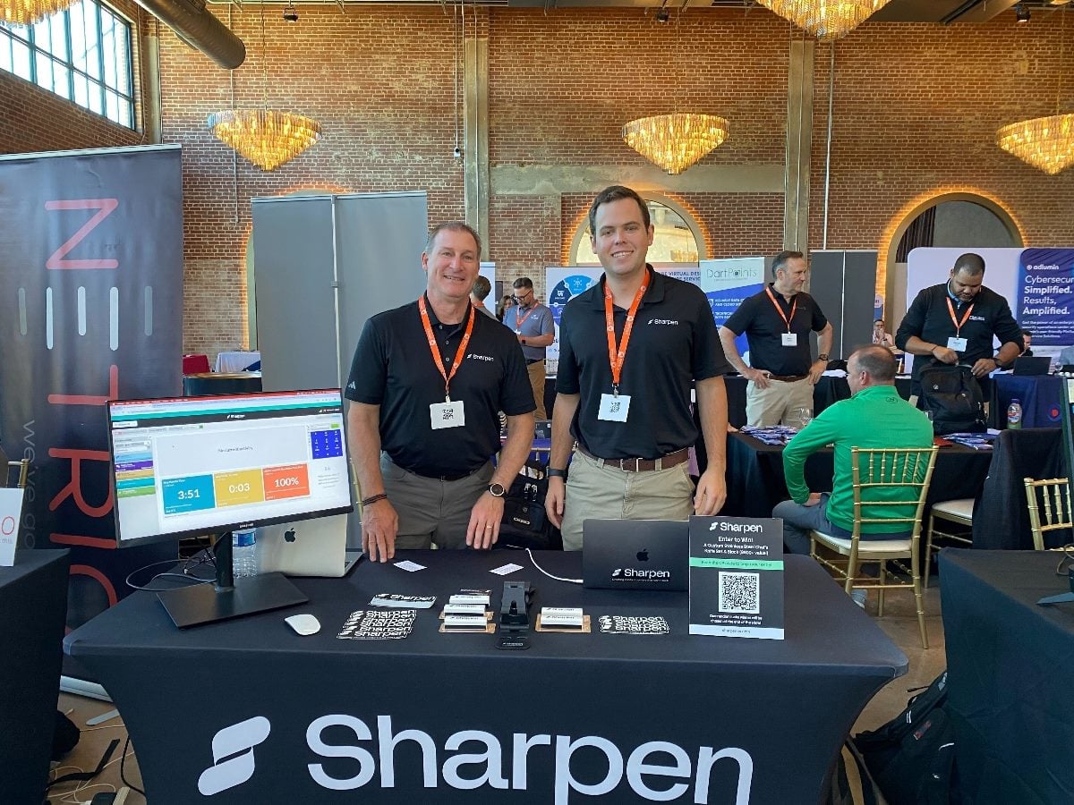 Two SharpenCX team members stand at their booth during the Opkalla Tech Summit in Charlotte, smiling and wearing branded black polos with orange lanyards. Their display table features laptops, product materials and a monitor demoing Sharpen’s cloud CCaaS platform as attendees network in the background inside a brick-and-glass event space with large chandeliers.