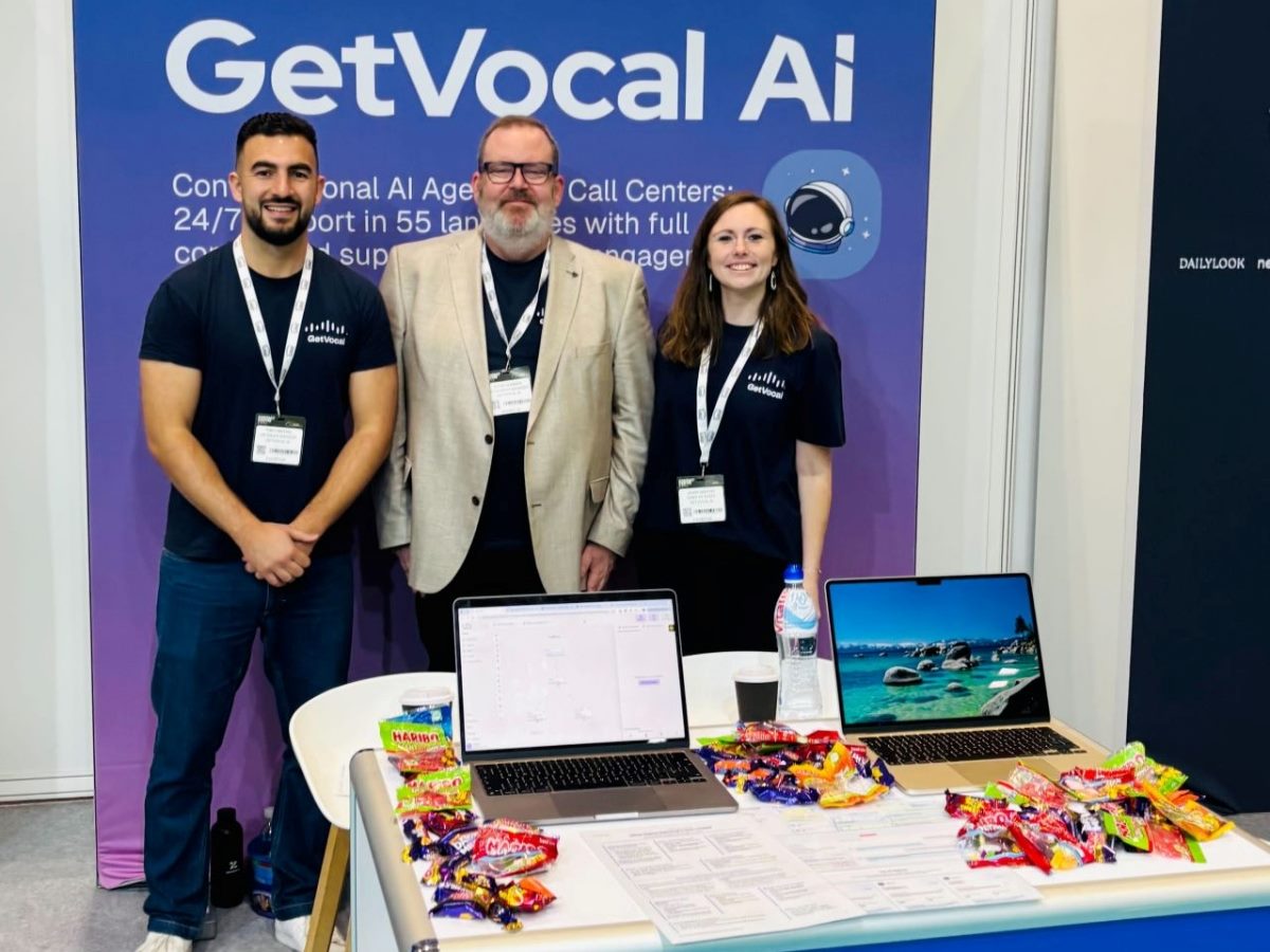 Three GetVocal AI team members stand smiling at their conference booth, with two laptops, printed materials and a large spread of assorted candy displayed on the table in front of them. A purple backdrop with the GetVocal AI logo and messaging appears behind them.