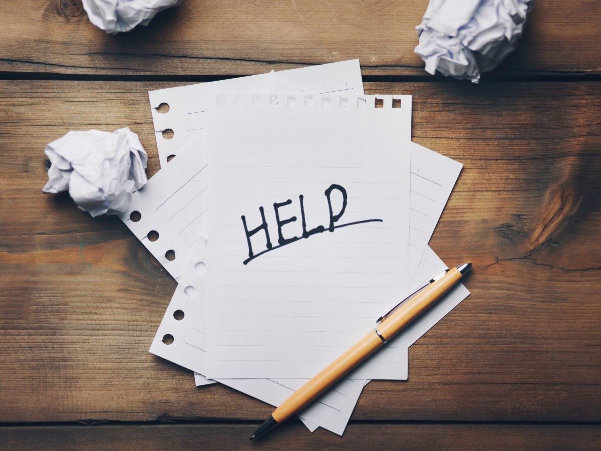 A sheet of paper with the word “HELP” handwritten in large black letters sits on a wooden desk surrounded by crumpled paper and a pen, symbolizing frustration, writer’s block or the need for support.