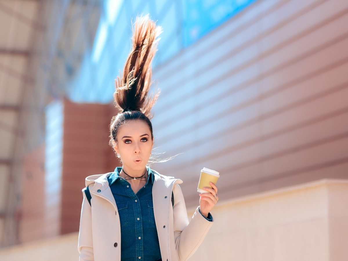 A surprised young woman with her hair flying straight up holds a takeaway coffee cup outdoors, standing against a modern building facade.