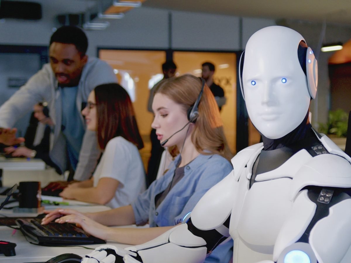 A humanoid robot wearing a headset sits beside human call center agents, symbolizing the collaboration between AI and people in customer support.