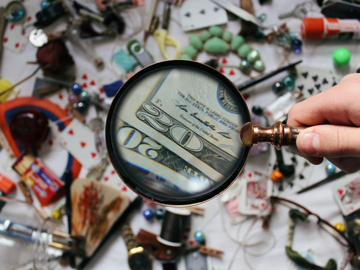 A person holds a magnifying glass focusing on a twenty-dollar bill among a cluttered table filled with various objects like playing cards, glue and small trinkets.