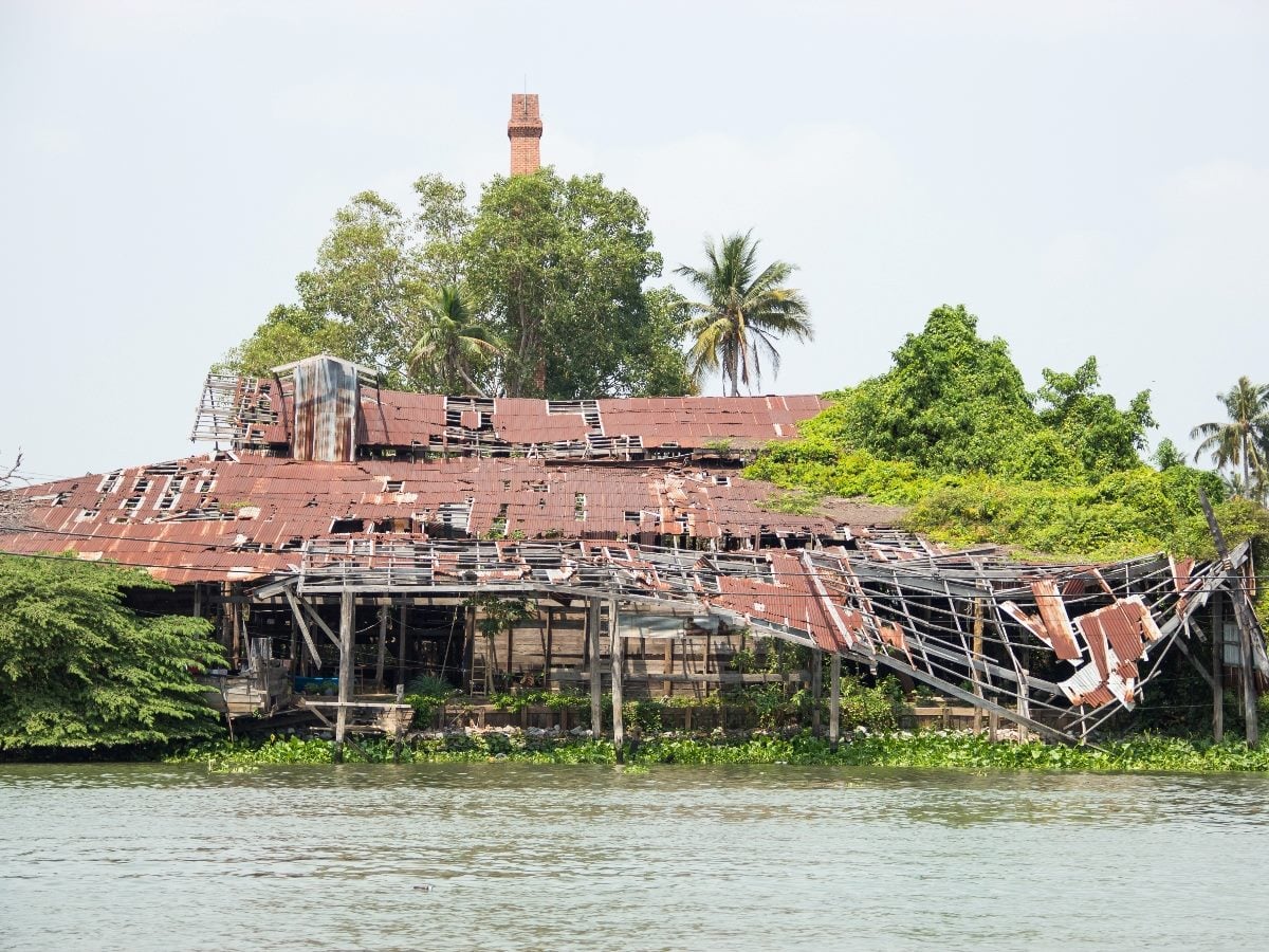 Dilapidated wooden building with a rusted, partially collapsed metal roof along a riverbank, surrounded by overgrown vegetation and palm trees.
