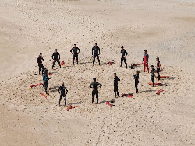 circle of people in wetsuits holding a meeting on a beach