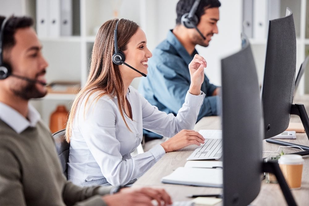 People working in a busy contact center sitting at a long table with computer screens in front of them and wearing headsets suggesting the need for a customer-centric approach.