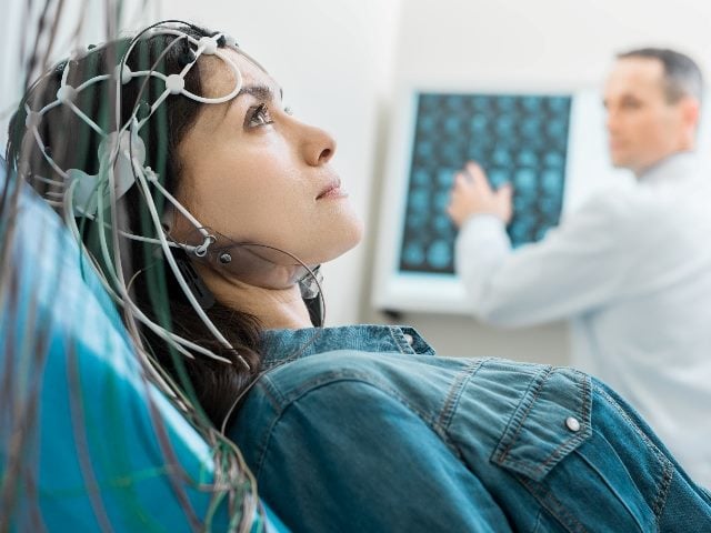 Young woman laying down undergoing electroencephalography as a doctor watches on in the background.