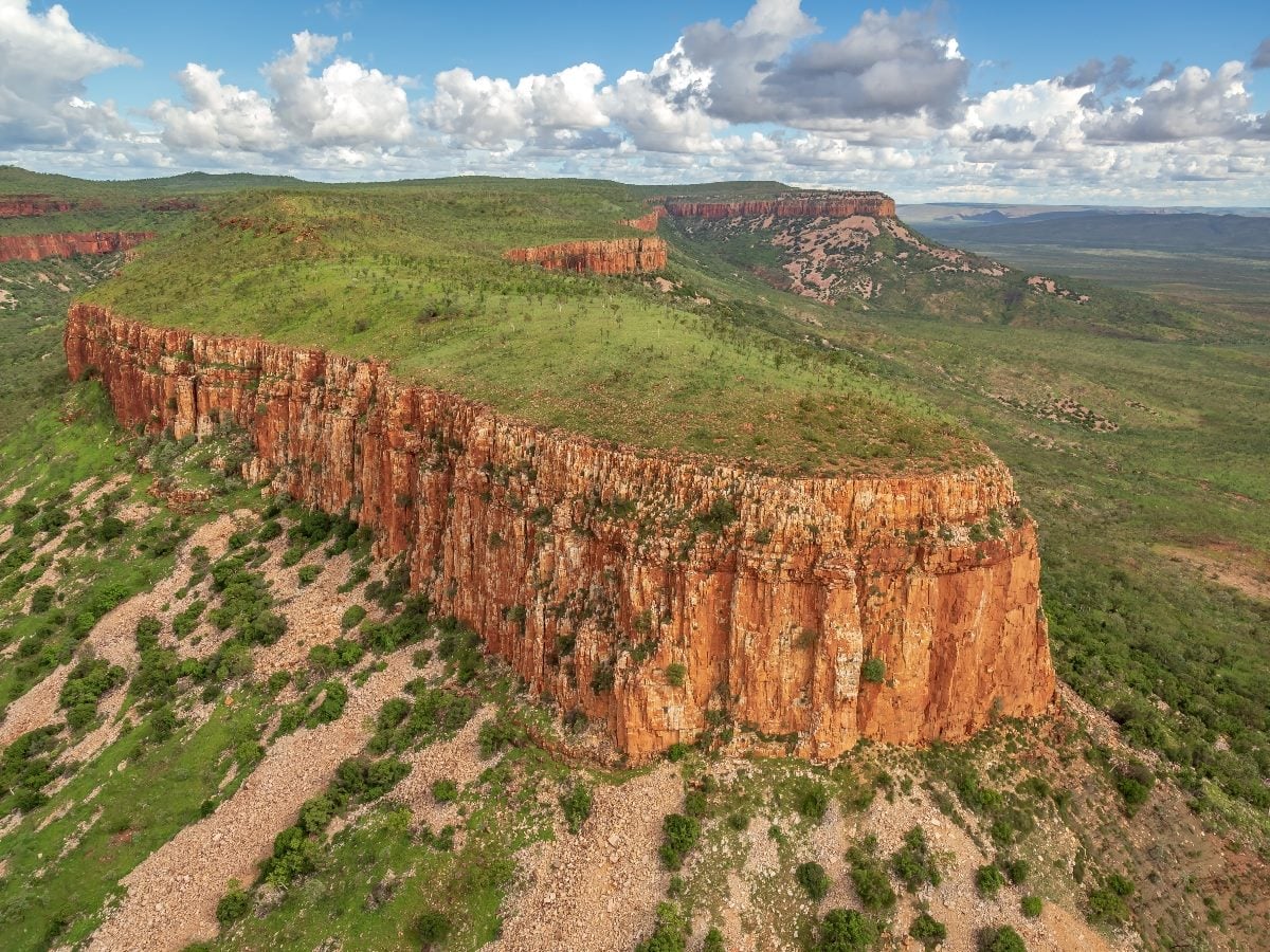 Aerial view of a wide, flat-topped plateau with steep red rock cliffs and scattered green vegetation, stretching into a vast landscape under a partly cloudy sky.