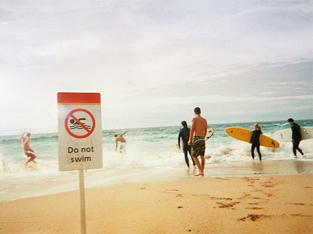 Several people in swimsuits and some with surfboards at the beach ignoring a "Do not swim" sign.