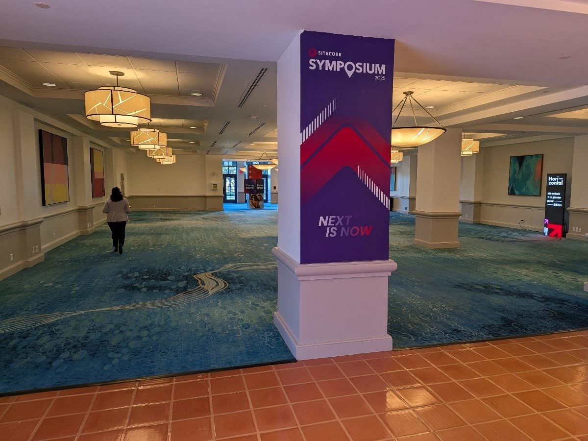 A hallway inside the Sitecore Symposium 2025 venue with a purple event sign on a column reading “NEXT IS NOW.” The carpet is patterned in shades of blue, and a person walks toward the distant lobby under warm ceiling lights.