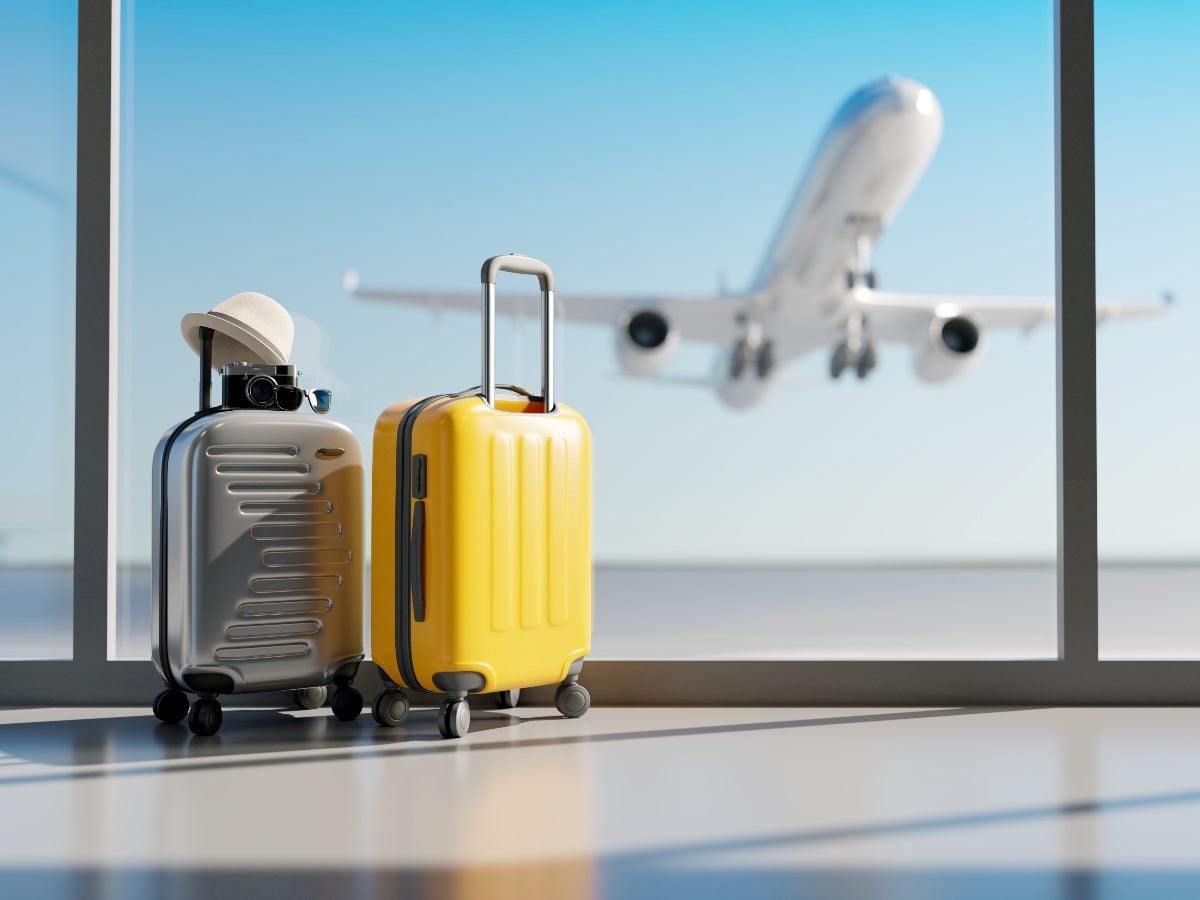 Two rolling suitcases sit near a large airport terminal window as a passenger jet takes off in the background. One suitcase is bright yellow, while the other is silver and topped with a hat and sunglasses, evoking leisure travel and vacation planning.