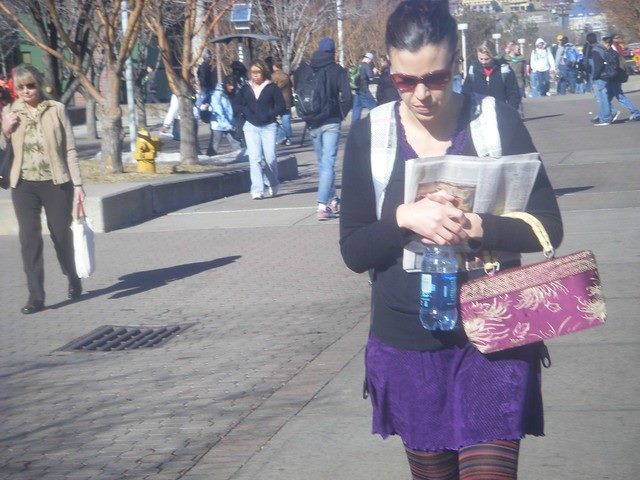 Woman holds a newspaper, a bag and a cup as she walks outside.