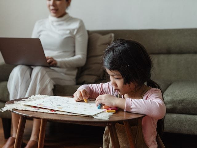 woman working on her couch while her child draws a few feet away