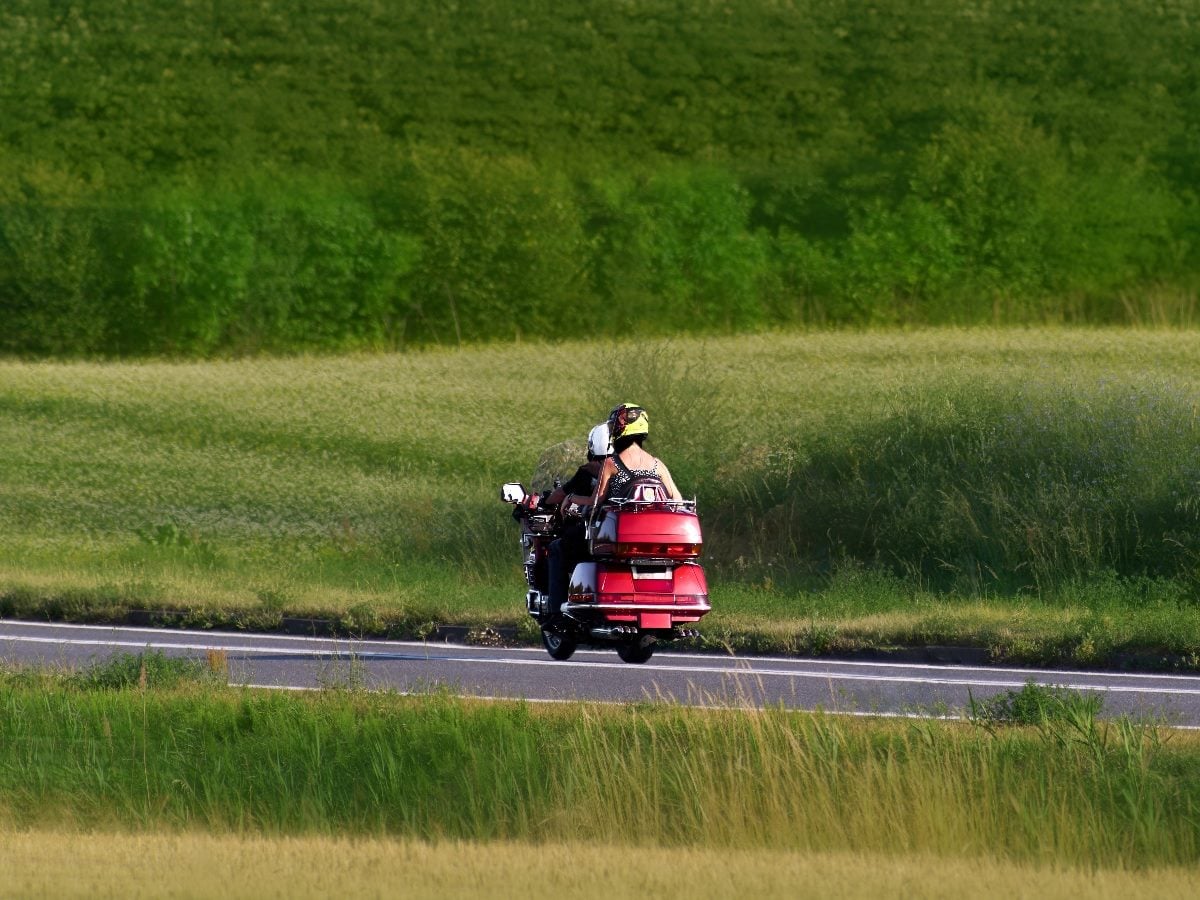 Two people ride a red touring motorcycle along a quiet rural road bordered by tall grass and green fields on a sunny day.