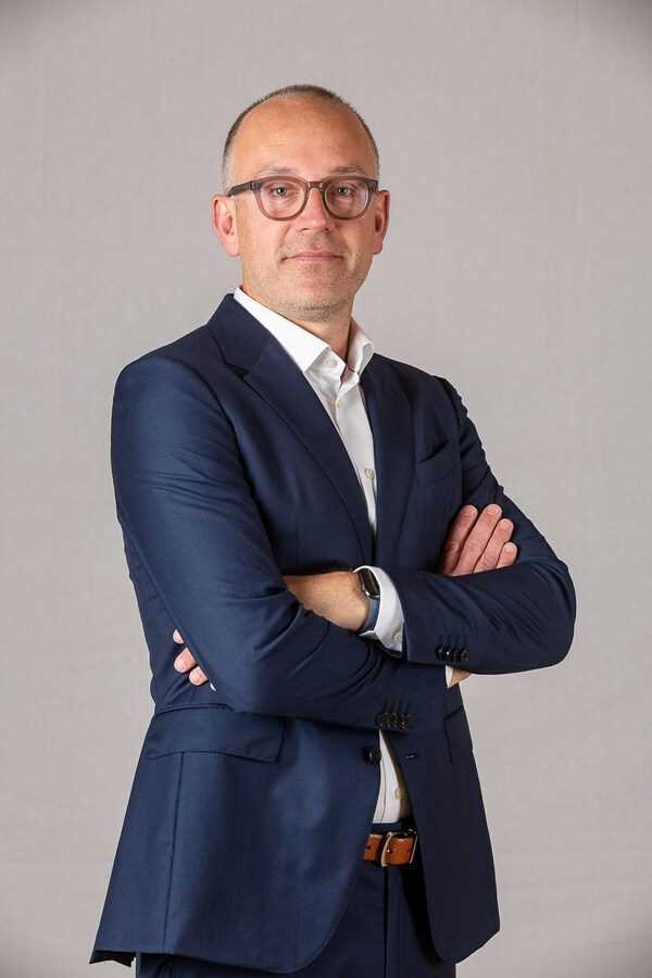 Dave O'Flanagan, who was recently appointed as Sitecore's interim CEO, stands with arms crossed and wears a blue suit and glasses in piece about Sitecore leadership.