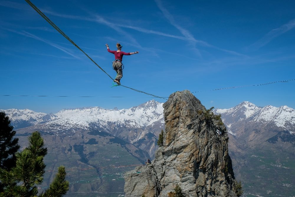 A person in a red top and beige pants is balancing on a slackline stretched between two rocky peaks, high above a scenic valley with a panoramic view of snowy mountains and clear blue skies in piece about economic strategies.