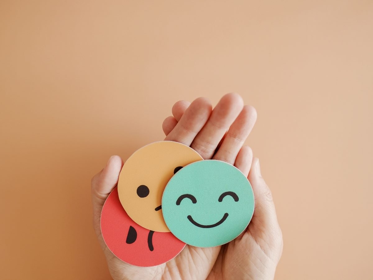 A hand holding three round emoji faces representing different emotions—sad (red), neutral (orange), and happy (green)—against a peach background.