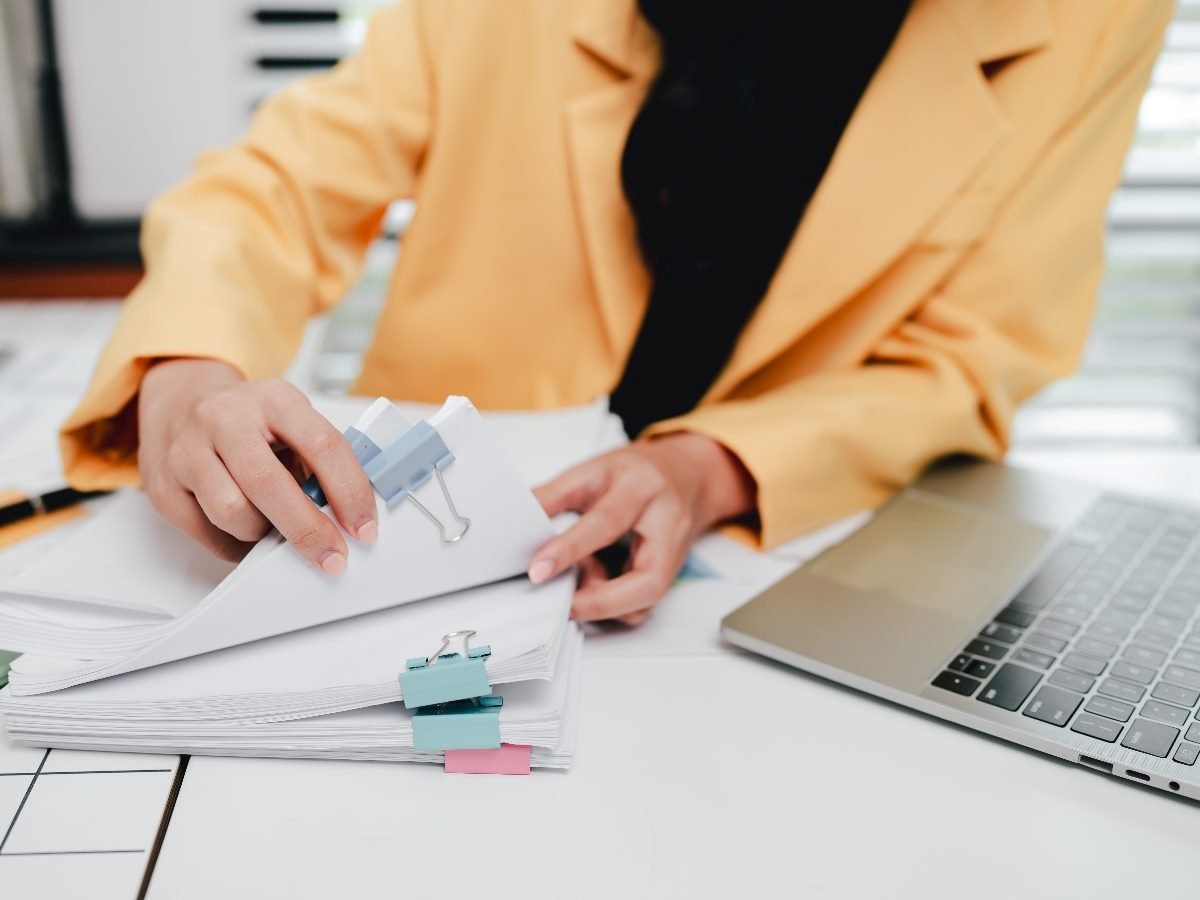A person in a yellow blazer reviews a stack of documents clipped together, with a laptop open on the desk beside them.