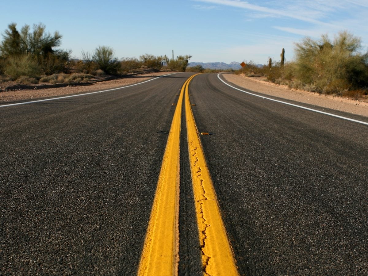 A two-lane asphalt highway stretches through a desert landscape, with bright yellow center lines leading toward a gentle curve in the road. Sparse desert vegetation and cacti line both sides of the road under a clear blue sky, with distant mountains visible on the horizon.