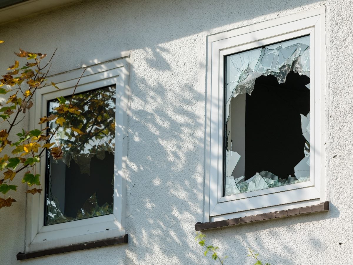 Broken windows on the exterior of a light-colored house, with shattered glass in both panes and jagged edges surrounding large holes; tree branches and leaves partially frame the scene.