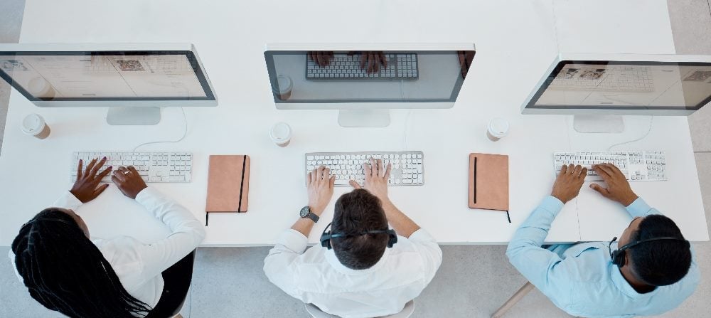 A top view of three employees wearing headsets and using computers working in a contact centers in piece about AI workplace integration.