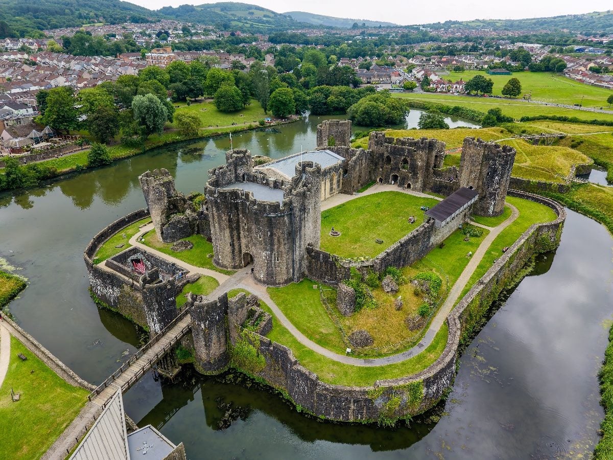 Aerial view of a historic stone castle surrounded by a wide moat and green grounds, with a bridge entrance and a town spreading across rolling hills in the background.