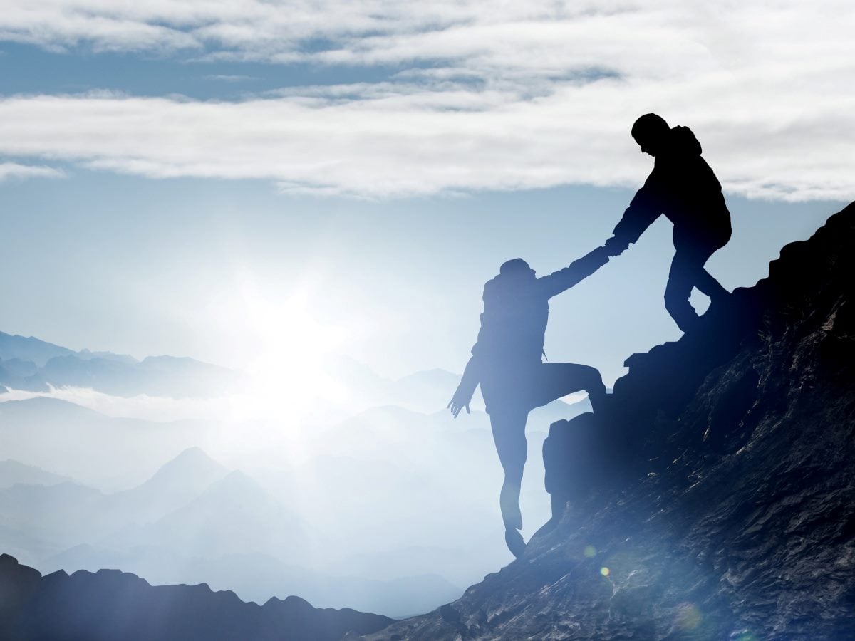 Two hikers in silhouette climb a steep rocky ridge at sunrise, with one climber positioned higher on the rocks reaching down to grasp the hand of a second climber below, who has one leg extended forward to step onto a higher foothold while the other leg pushes off a lower rock. Both wear backpacks and jackets, rendered as dark silhouettes against a hazy blue sky. The sun sits low near the horizon, casting a bright glow and lens flare through thin clouds. Behind them, layered mountain ridges fade into pale blue mist toward the horizon, receding in depth from darker foreground peaks to nearly white distant ranges. The overall palette is cool blue-gray, evoking early morning light and high altitude.