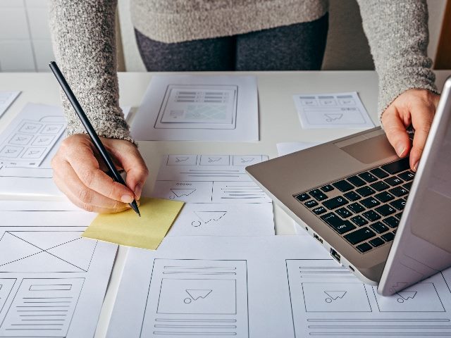 A web designer wearing a sweater and  working on laptop and website wireframe sketches on white table in piece about website redesign. 