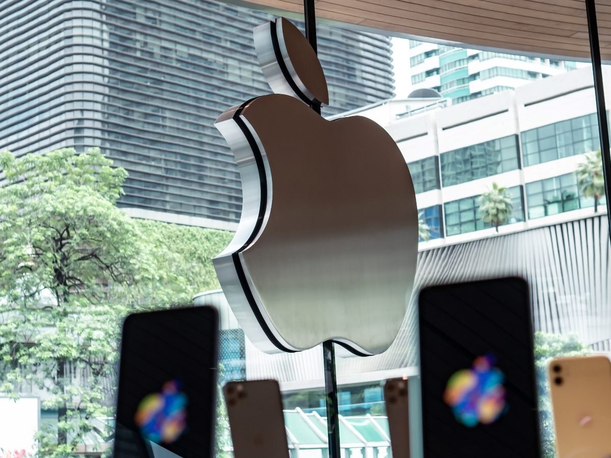 The Apple logo displayed outside a company retail store with modern city buildings and trees in the background.