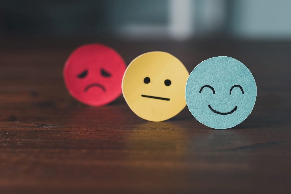 A red sad face, a yellow neutral face and a blue happy face are displayed on paper cutouts with faces drawn in black marker in piece about empathy in customer service and emotional intelligence.