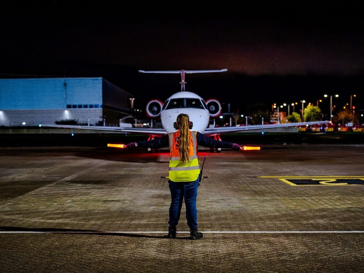 Ground crew member wearing a high-visibility safety vest and holding illuminated marshalling wands directs a private jet on an airport tarmac at night. The aircraft faces the camera as runway and terminal lights glow in the background, highlighting the coordination and signaling required to guide aircraft safely on the ground.