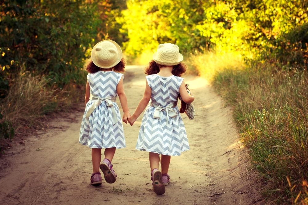 Back view of two little girl twins wearing identical gray and white striped dresses and straw hats while holding hands in piece about digital twins.