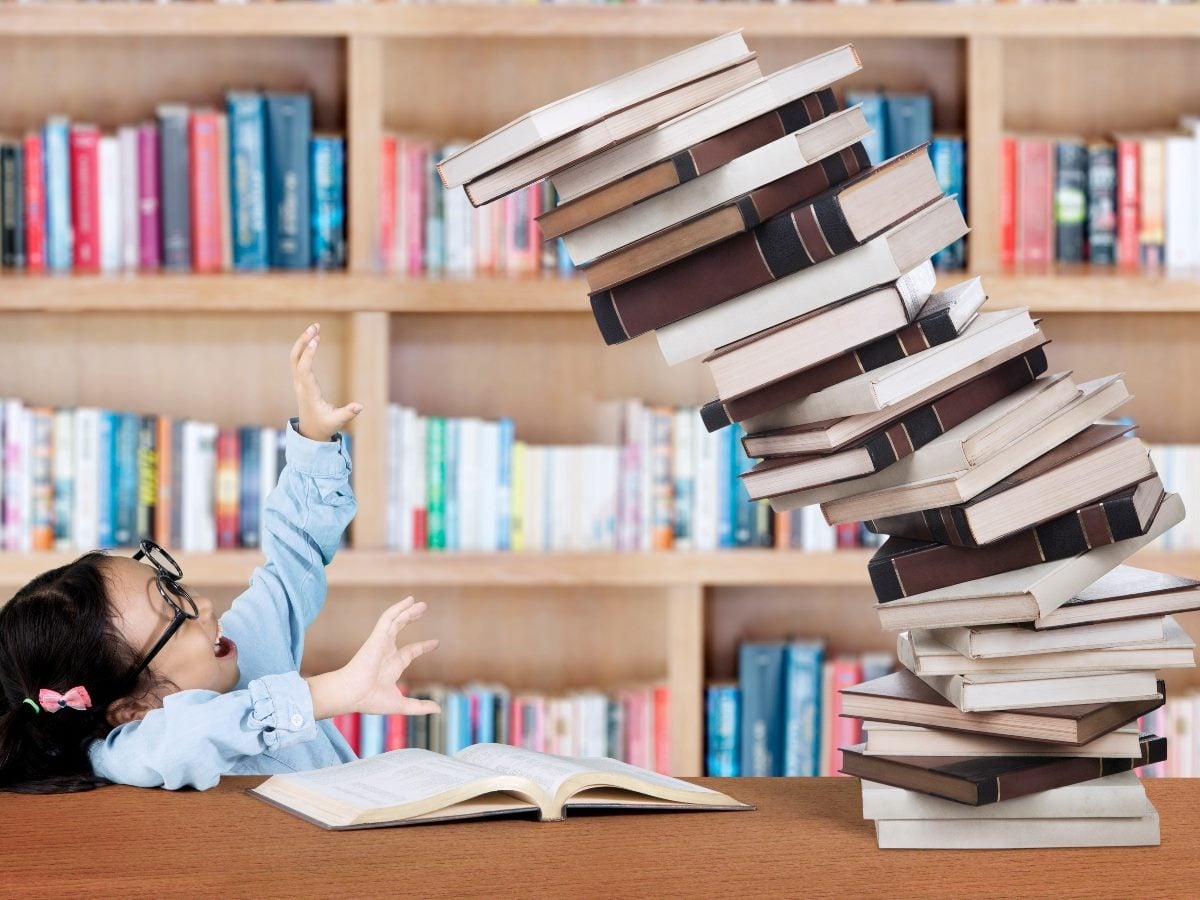 Child in a library reaching toward a dangerously leaning stack of books beside an open book on a table.