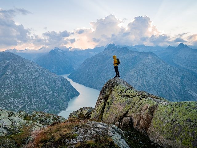 Hiker standing on top of a rocky mountain overlooking the dramatic landscape at sunset at Grimselsee Lake, Switzerland. 